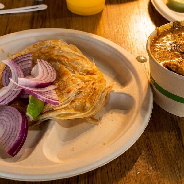 Flaky paratha with red onions and lime on a compostable plate, beside a curry cup.