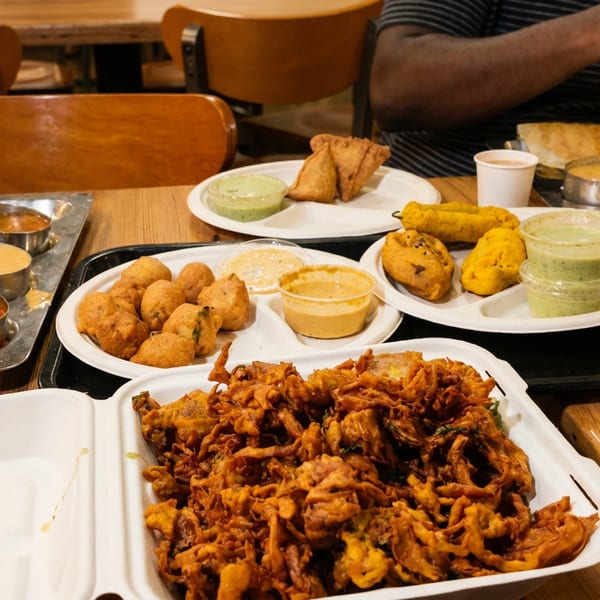 Table spread with various Indian dishes like dosa, pakoras, samosas, and chutneys, with diners.