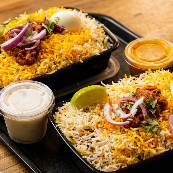 Two biryani meals with rice, meat, boiled egg, lime, and red onions on a black tray.
