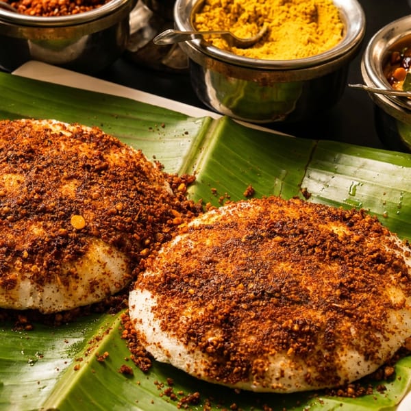 Two spice-dusted idlis on banana leaves with various chutneys and powders in the background.