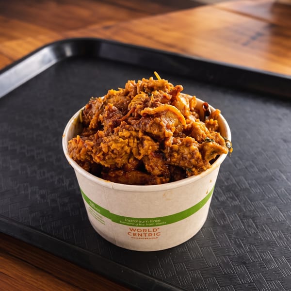 A close-up of a paper cup filled with saucy, seasoned chicken pieces on a black tray.